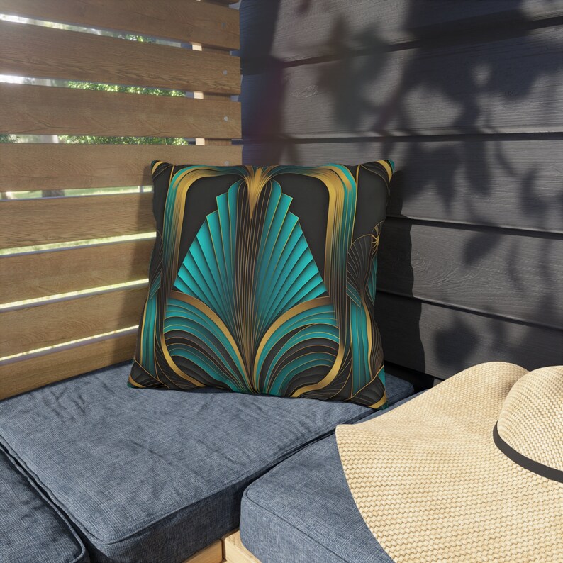 May include: Teal and gold art deco patterned throw pillow. The pillow features a geometric design with teal and gold accents on a black background.  The pillow is shown on a dark blue outdoor couch with a beige sunhat resting on the armrest. The pillow is square-shaped and appears to be made of a soft, durable fabric suitable for outdoor use. The design is reminiscent of 1920s and 1930s art deco style. The pillow would add a touch of elegance and sophistication to any outdoor space.