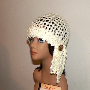 May include: Cream-colored crochet hat with a decorative button and a flowing, fringed side detail. The hat is displayed on a mannequin head, showcasing its open weave design and textured brim. A vintage-inspired accessory.