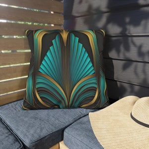 May include: Teal, gold, and black art deco-style throw pillow. The pillow features a geometric design with teal and gold accents on a black background.  The design is reminiscent of Art Deco architecture and features repeating patterns. The pillow is shown on a blue outdoor furniture cushion.