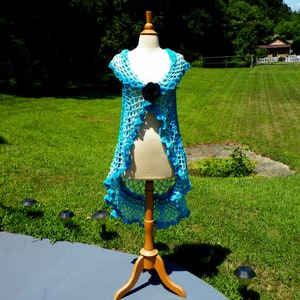 May include: A handmade, open-front crochet vest in shades of blue, displayed on a wooden dress form. The vest features a scalloped edge and a black flower accent. The garment is displayed outdoors on a sunny day.