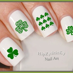 Peut inclure: Autocollants pour ongles en forme de trèfle vert. Les autocollants présentent différents motifs de trèfles, notamment un trèfle unique, un groupe de trèfles et un trèfle avec un motif en spirale. Les autocollants sont sur un vernis à ongles blanc.