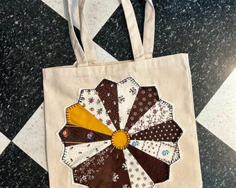 Könnte beinhalten: Beige Canvas-Tragetasche mit einem großen, farbenfrohen Patchwork-Design. Das Design zeigt ein kreisförmiges Muster mit braunen, weißen und gelben Stoffstücken. Die Tasche hat zwei beige Griffe.
