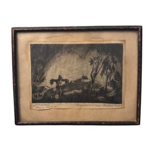 Op de afbeelding: Ingelijst zwart-wit kunstwerk met een figuur die een kruis draagt, geplaatst in een landschap met bomen en een gebouw. Het kunstwerk zit in een donkere houten lijst met een lichtgekleurde passe-partout. Tekst is zichtbaar aan de onderkant.