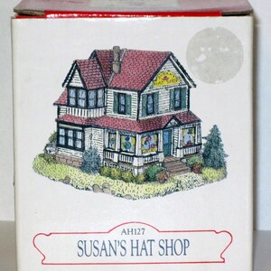 Peut inclure: Une boîte blanche avec un dessus rouge et un dessin d'une maison blanche avec un toit rouge. La maison a une enseigne qui dit "Susan's Hat Shop". La boîte porte le texte "AH127 Susan's Hat Shop" en bas.