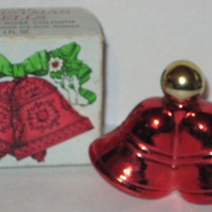 May include: A red glass bottle shaped like two bells with a gold top. The bottle is sitting on a white surface. The bottle is labelled "Christmas Bells" and "1 fl oz".