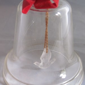 May include: A clear glass bell ornament with a white dove inside. The bell has a red velvet bow on top and the year 1983 is etched on the bottom.