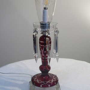 May include: A red glass table lamp with a clear glass shade and crystal prisms hanging from the base. The lamp has a white cord and a clear glass base.