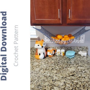 May include: A grey crocheted hammock hanging from kitchen cabinets, filled with orange mandarins. The text "Stitched by JoAnna" is visible below the hammock. The counter below the hammock has a variety of decorative items, including a blue waffle maker, a white ceramic fox figurine, and a white ceramic pig figurine.