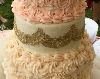 Puede incluir: Un pastel de boda de tres pisos decorado con delicado glaseado de crema de mantequilla rosa y blanco. Los dos pisos inferiores presentan un diseño con volantes, mientras que el piso superior está adornado con un borde de encaje dorado. El pastel está coronado con un adorno de pastel dorado.