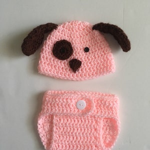 May include: A light pink crocheted baby hat and diaper set with brown ears and a brown nose. The hat has two black buttons for eyes.