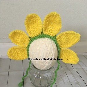 May include: A crocheted sunflower headband with yellow petals and green leaves. The headband is displayed on a white yarn ball and a glass jar. The text "HandcraftedWitches" is visible on the image.