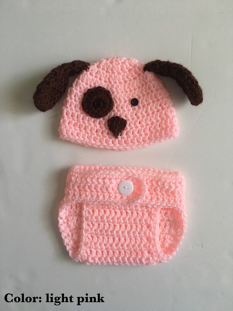 May include: A light pink crocheted baby hat and diaper set with brown ears and a brown nose. The hat has two black buttons for eyes.