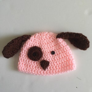 May include: A light pink crocheted baby hat with brown ears and a brown face. The hat has two black buttons for eyes and a brown nose.