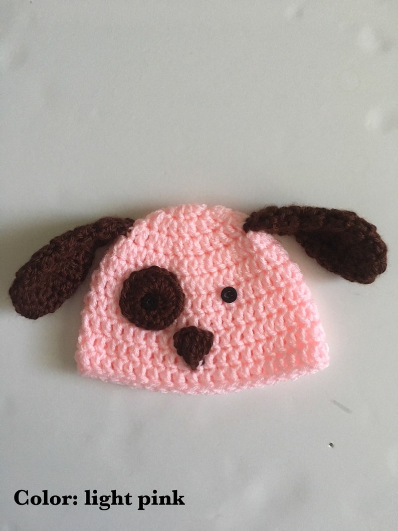 May include: A light pink crocheted baby hat with brown ears and a brown face. The hat has two black buttons for eyes and a brown nose.