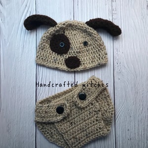May include: A crocheted dog hat and diaper set in beige and brown with black buttons. The hat has brown ears and a brown nose with blue eyes. The diaper has two black buttons. The set is handcrafted.