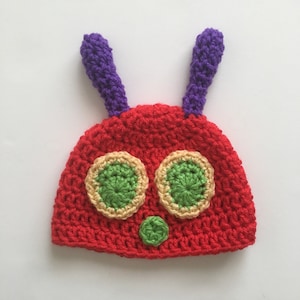 Peut inclure: Un bonnet rouge en crochet avec des yeux verts et jaunes et un nez vert. Le bonnet a deux antennes violettes.