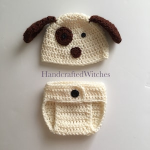 May include: A crocheted baby outfit in light cream with a dog hat featuring brown ears and a brown nose. The hat has a brown button for an eye and a brown button for a nose. The outfit includes a diaper cover with a brown button.