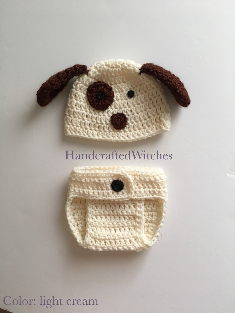 May include: A crocheted baby outfit in light cream with a dog hat featuring brown ears and a brown nose. The hat has a brown button for an eye and a brown button for a nose. The outfit includes a diaper cover with a brown button.