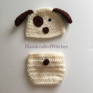 May include: A crocheted dog hat and diaper set in light cream with brown accents. The hat has floppy ears and a brown nose and eyes. The diaper has a brown button on the front. HandcraftedWitches.