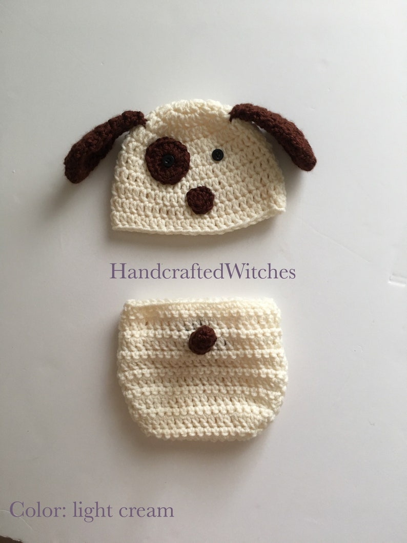 May include: A crocheted dog hat and diaper set in light cream with brown accents. The hat has floppy ears and a brown nose and eyes. The diaper has a brown button on the front. HandcraftedWitches.