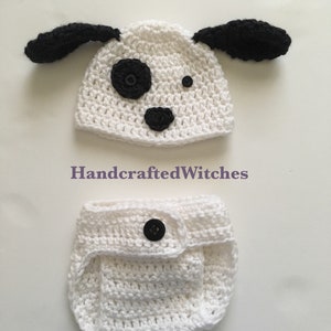 May include: White crocheted baby hat and diaper set with black ears, nose, and eyes. The hat has a black button for a nose and a black button for an eye. The diaper has a black button on the front.