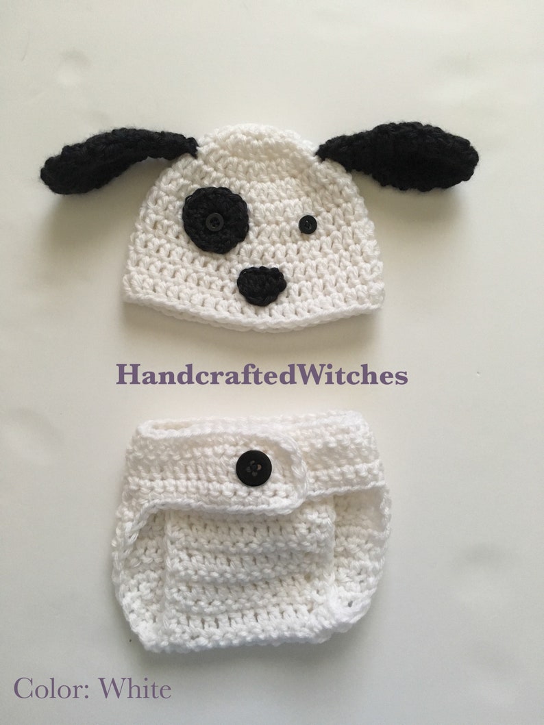 May include: White crocheted baby hat and diaper set with black ears, nose, and eyes. The hat has a black button for a nose and a black button for an eye. The diaper has a black button on the front.
