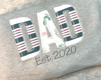 May include: Gray sweatshirt with the word "DAD" in white fabric with red and black stripes. The letters are sewn on with a white outline. The text "Est. 2020" is embroidered in gray below the word "DAD".