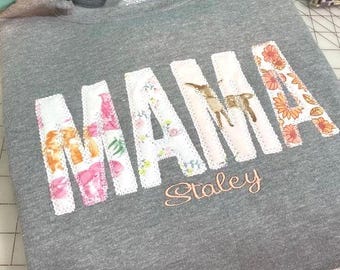 May include: A gray sweatshirt with the word "MAMA" spelled out in large, white letters. Each letter is made of fabric scraps sewn together in a patchwork style with floral and animal prints. The name "Staley" is embroidered in orange below the word "MAMA".