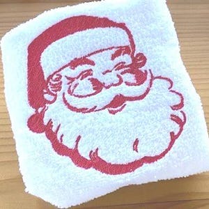 May include: White towel with a red embroidered image of Santa Claus's face. The embroidery is detailed and includes Santa's beard, mustache, and hat.