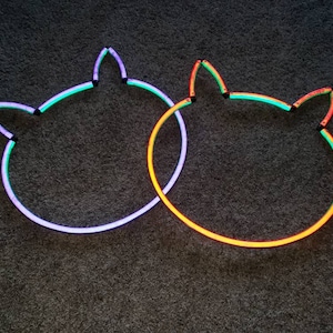 May include: Two neon cat head outlines on a dark gray background. The outline on the left is purple, green, and blue. The outline on the right is orange, green, and yellow.
