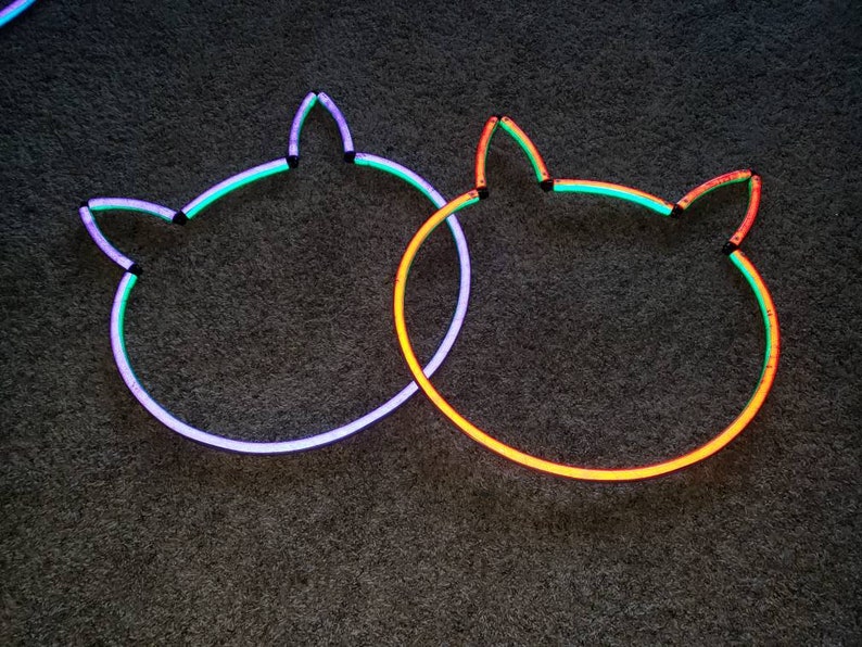 May include: Two neon cat head outlines on a dark gray background. The outline on the left is purple, green, and blue. The outline on the right is orange, green, and yellow.