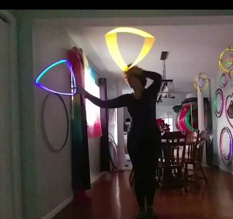 K&ouml;nnte beinhalten: Eine Person f&uuml;hrt eine Performance mit zwei LED-Hula-Hoops durch. Die Reifen leuchten in verschiedenen Farben und erzeugen ein farbenfrohes Schauspiel. Die Person tr&auml;gt schwarze Kleidung und befindet sich in einem Raum mit Holzboden. Im Hintergrund h&auml;ngen weitere Reifen an der Wand.