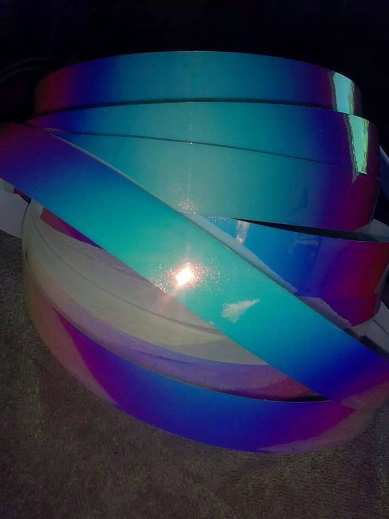 Puede incluir: Un rollo de vinilo iridiscente en tonos azules, morados y rosas. El vinilo tiene un acabado hologr&aacute;fico que brilla a la luz.