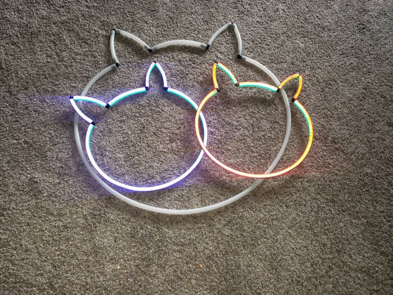May include: Two neon cat head outlines, one blue and green, the other orange and yellow, are intertwined on a gray carpet.