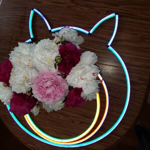 May include: A bouquet of white and pink peonies and red roses arranged inside a neon light shaped like a cat's head.