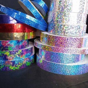 May include: Assortment of iridescent holographic ribbon rolls in various colors, including blue, purple, red, gold, and silver. The rolls are stacked on top of each other.