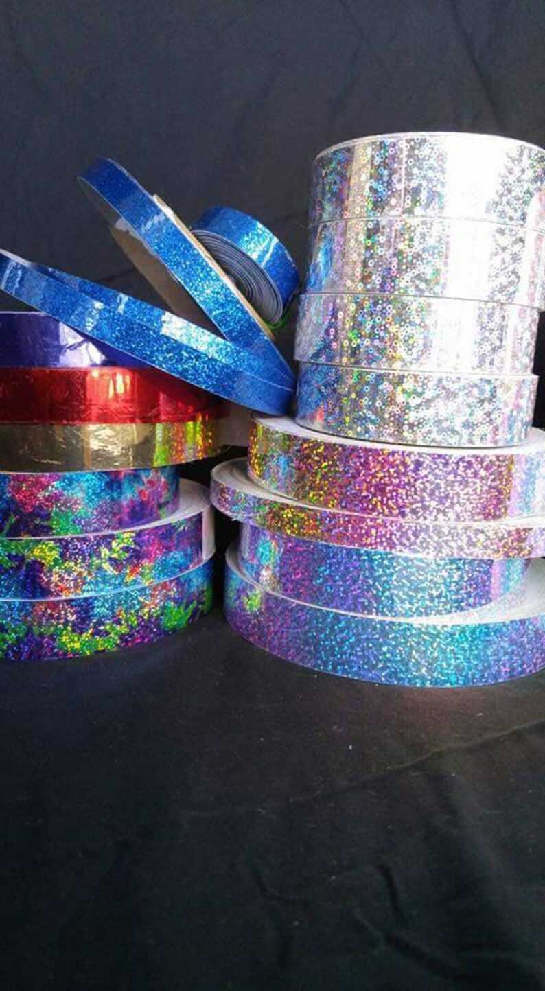 May include: Assortment of iridescent holographic ribbon rolls in various colors, including blue, purple, red, gold, and silver. The rolls are stacked on top of each other.