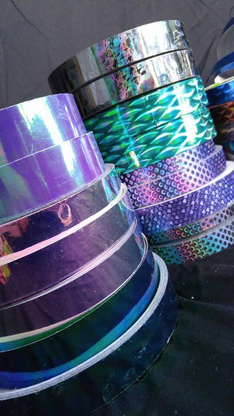 May include: A stack of iridescent holographic tape rolls in various colors, including purple, blue, green, and black. The tape is perfect for crafting, decorating, and adding a touch of sparkle to any project.