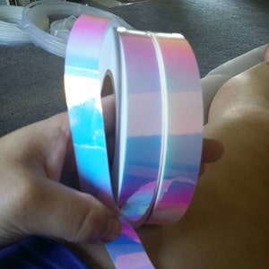 Puede incluir: Dos rollos de cinta hologr&aacute;fica iridiscente con un n&uacute;cleo blanco. La cinta es un degradado de rosa, azul y morado.