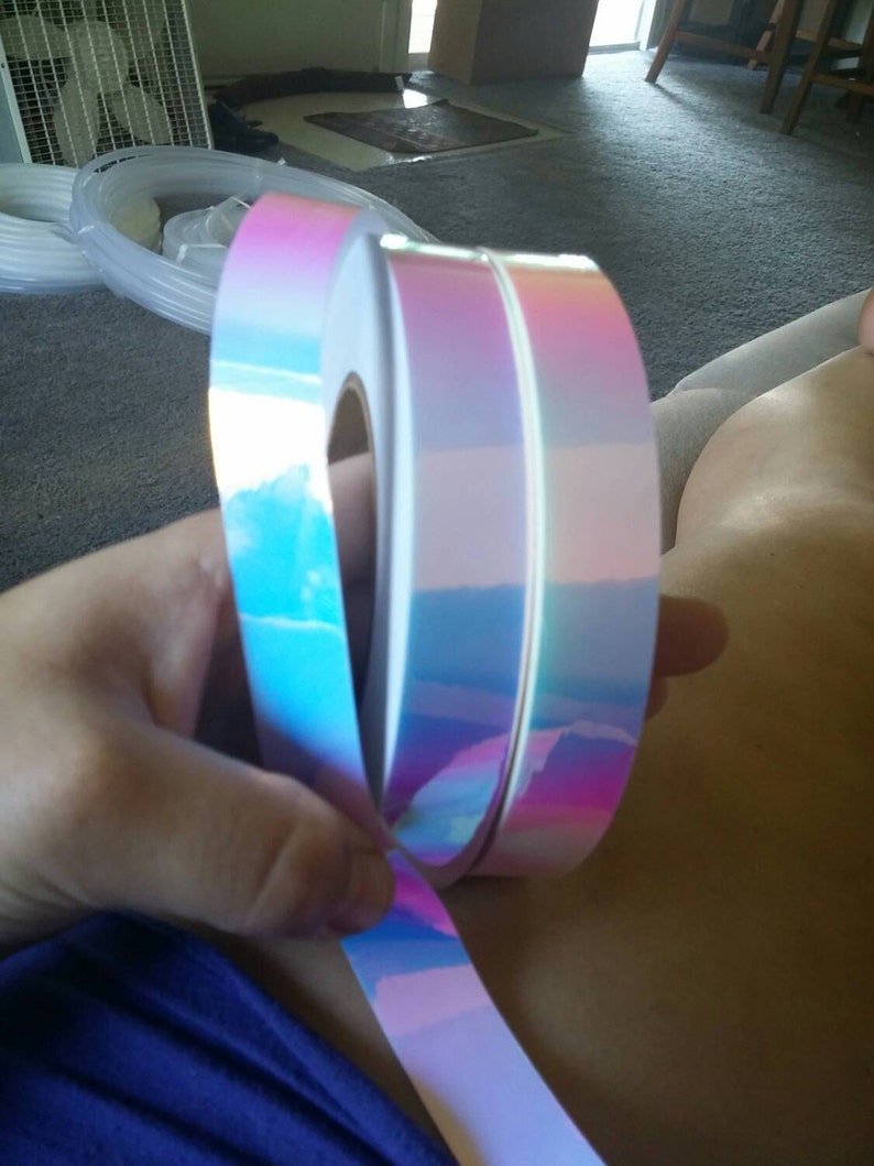 Puede incluir: Dos rollos de cinta hologr&aacute;fica iridiscente con un n&uacute;cleo blanco. La cinta es un degradado de rosa, azul y morado.