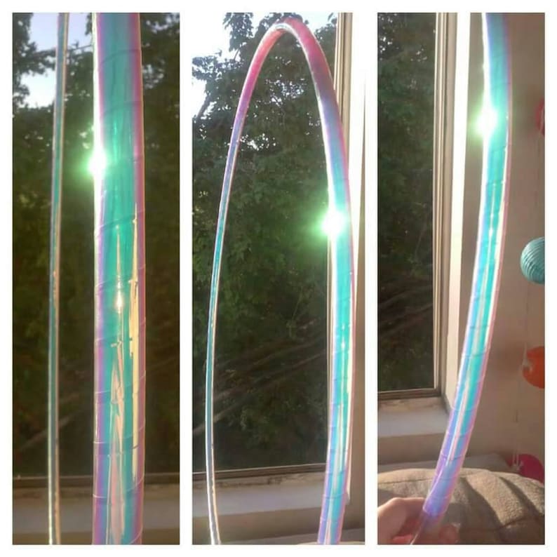 Puede incluir: Un aro grande iridiscente con un esquema de colores azul, verde y rosa. El aro est&aacute; hecho de pl&aacute;stico y es flexible. Est&aacute; siendo sostenido por una mano frente a una ventana.