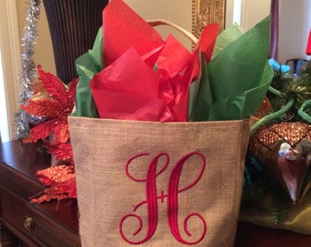 Puede incluir: Una bolsa de regalo de arpillera con una letra H bordada en rojo. La bolsa está llena de papel de seda rojo y verde.
