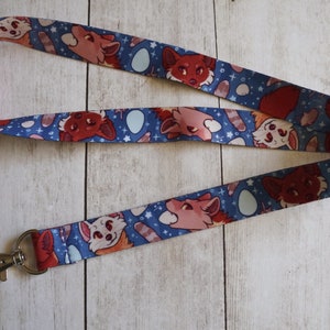 May include: A blue lanyard with a white background featuring a repeating pattern of red pandas with blue and white eggs. The lanyard has a silver metal clip.