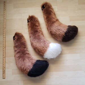 May include: Three fluffy fox tails in different sizes. The tails are brown with black tips and one tail has a white tip.