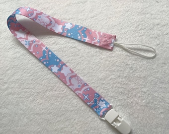Puede incluir: Un clip para chupete con estampado rosa y azul con un clip de plástico blanco. El clip tiene una cuerda blanca.