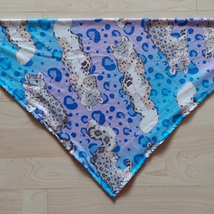 K&ouml;nnte beinhalten: Ein dreieckiger Schal mit einem blauen und violetten Farbverlauf als Hintergrund. Der Schal ist mit einem sich wiederholenden Muster aus wei&szlig;en Schneeleoparden und blauen Punkten verziert.