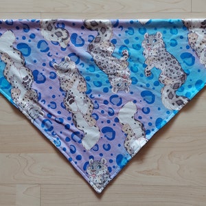 K&ouml;nnte beinhalten: Ein dreieckiges Bandana mit einem blauen und violetten Ombre-Hintergrund. Das Bandana ist mit einem sich wiederholenden Muster aus wei&szlig;en Schneeleoparden und blauen Punkten bedeckt.