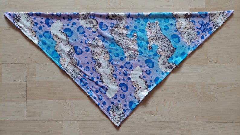 K&ouml;nnte beinhalten: Ein dreieckiges Bandana mit einem blauen und violetten Ombre-Hintergrund. Das Bandana ist mit einem sich wiederholenden Muster aus wei&szlig;en Schneeleoparden und blauen Punkten bedeckt.