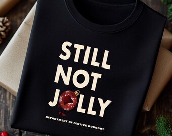 May include: Black sweatshirt with the text "STILL NOT JOLLY" in white, with a red and white ornament graphic. The phrase "DEPARTMENT OF FESTIVE BURNOUT" is printed below the main text.