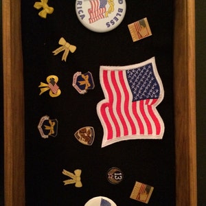 May include: A wooden shadow box with a variety of patriotic themed items, including a large American flag patch, a button that says "God Bless America", a button that says "I Support Our Troops", and several yellow ribbon pins.